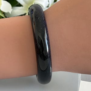 May include: A smooth, rounded bangle bracelet crafted from dark blue sandstone, featuring a polished finish with embedded glittery particles that create a starry, reflective appearance across the entire surface of the circular jewelry piece.