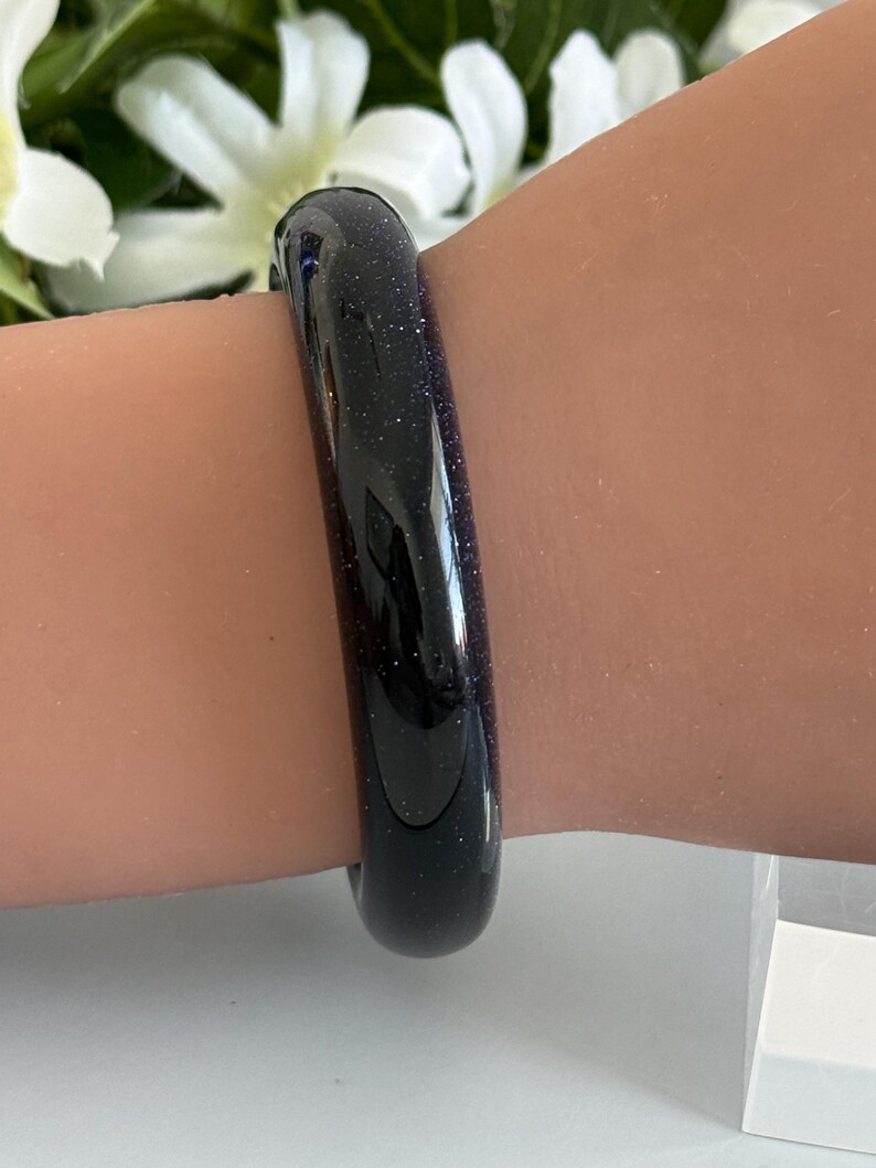 May include: A smooth, rounded bangle bracelet crafted from dark blue sandstone, featuring a polished finish with embedded glittery particles that create a starry, reflective appearance across the entire surface of the circular jewelry piece.
