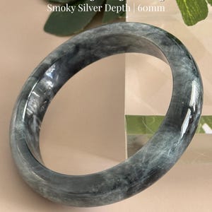 May include: A black and gray bangle with a smoky silver depth. The text on the image reads "Black Eagle Eye Bangle" and "Smoky Silver Depth | 60mm". The bangle is a smooth, circular shape, and the color is a mix of black and gray.