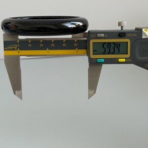 May include: A dark blue, glittery bangle bracelet measured by a digital caliper. The display shows a measurement of 59.34 mm. The bangle has a smooth, rounded surface with fine, sparkling inclusions throughout the deep blue material.
