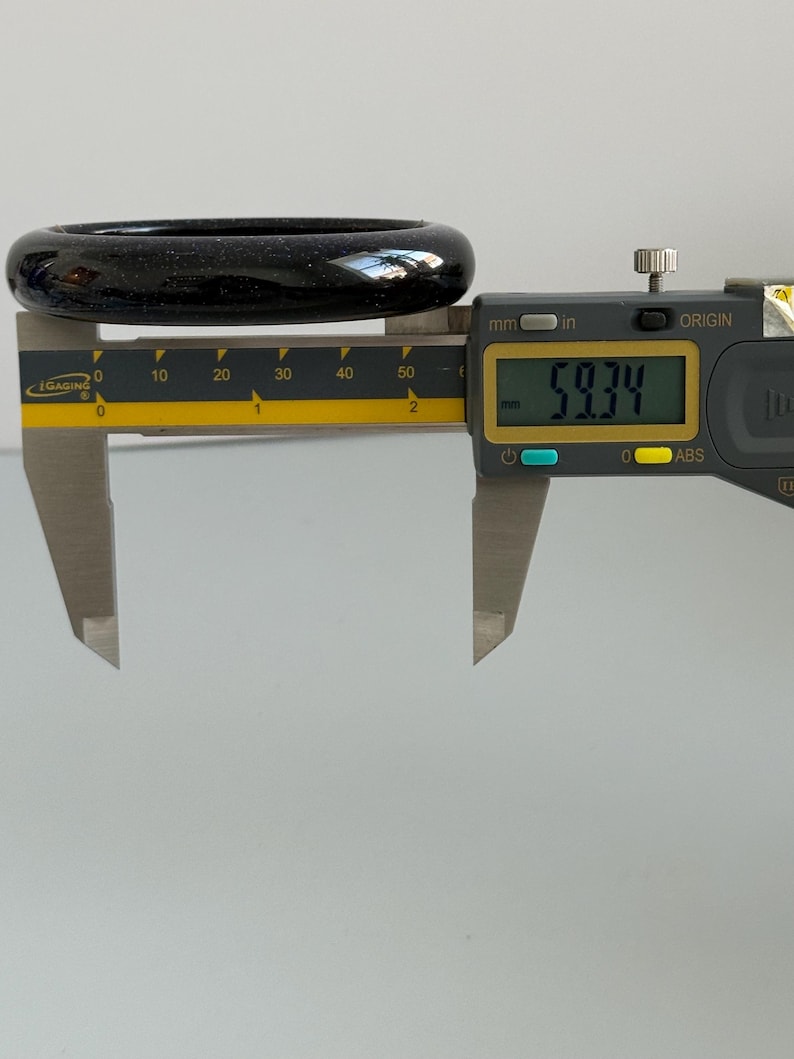 May include: A dark blue, glittery bangle bracelet measured by a digital caliper. The display shows a measurement of 59.34 mm. The bangle has a smooth, rounded surface with fine, sparkling inclusions throughout the deep blue material.