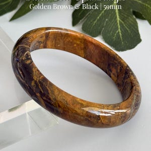 May include: A polished bangle bracelet featuring a marbled pattern of golden brown and black colors. The text on the image reads Black Gold Opal Bangle, Golden Brown & Black | 59mm, and AmorGemsBoutique. The bangle has a smooth, rounded surface.