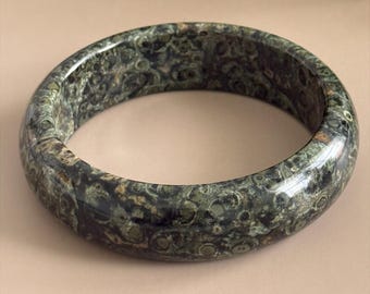 Può includere: Un bracciale rigido in diaspro Kambaba lucido con un motivo orbicolare verde scuro e nero. Il testo Kambaba Jasper Bangle Orbicular Beauty | 61.8 mm e AmorGemsBoutique è visibile. Il bracciale ha una superficie liscia e arrotondata di forma circolare.