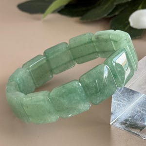 Pu&ograve; includere: Un braccialetto di avventurina verde. Il braccialetto &egrave; composto da pietre verdi rettangolari e lucide. Le pietre sono collegate per formare una fascia flessibile. Il braccialetto &egrave; mostrato su uno sfondo neutro con vegetazione.
