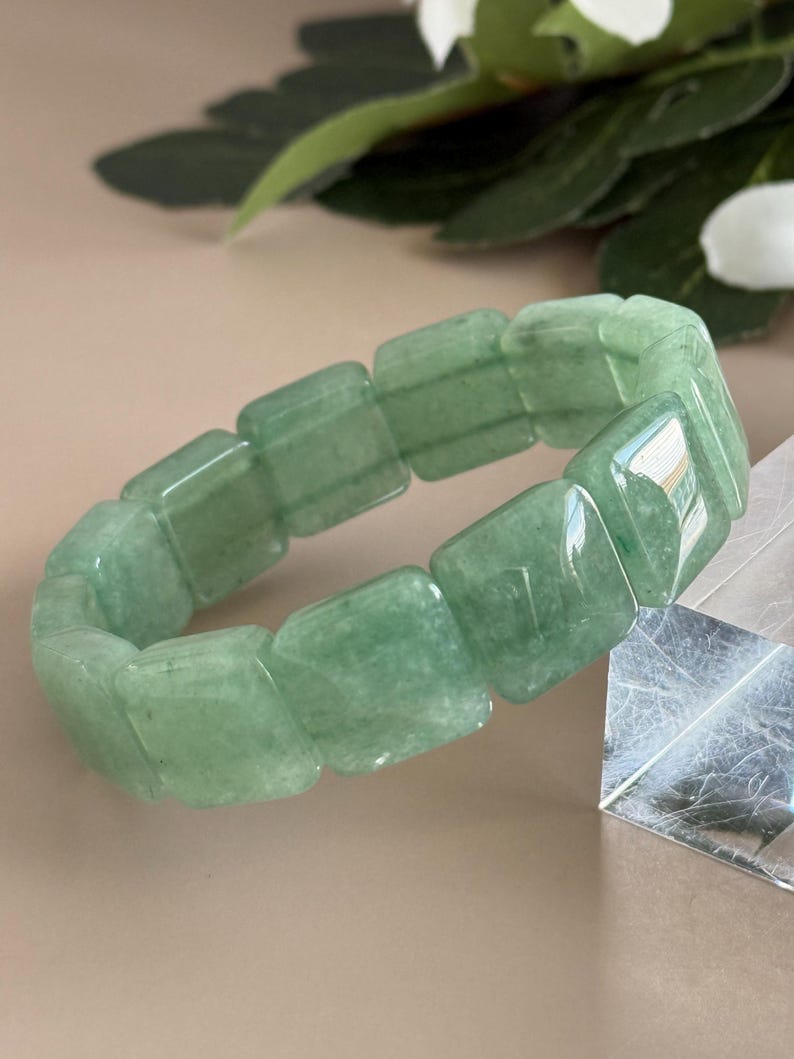 Pu&ograve; includere: Un braccialetto di avventurina verde. Il braccialetto &egrave; composto da pietre verdi rettangolari e lucide. Le pietre sono collegate per formare una fascia flessibile. Il braccialetto &egrave; mostrato su uno sfondo neutro con vegetazione.
