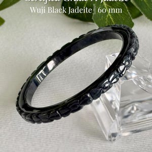 May include: A black Wuji jadeite bangle bracelet with a carved floral pattern along the outer edge. The text reads Certified Grade A Jadeite, Wuji Black Jadeite | 60 mm, and AmorGemsBoutique. The bangle has a smooth, polished finish.