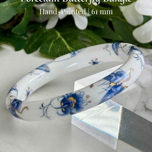 May include: A white porcelain bangle with hand-painted blue and gold butterfly and floral designs. The bangle is 61 mm in diameter.