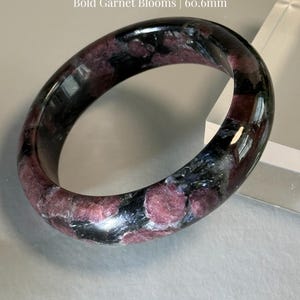 May include: A Firework Garnet bracelet with Bold Garnet Blooms. The polished gemstone bracelet is a deep black with red and white inclusions. The text "Firework Garnet" and "Bold Garnet Blooms | 60.6mm" are visible.