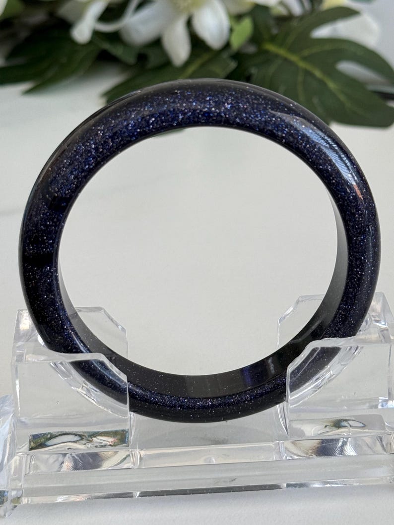 May include: A circular bangle bracelet crafted from dark blue sandstone, featuring a smooth, polished surface embedded with numerous tiny, reflective metallic flecks that create a shimmering, star-like appearance throughout the deep blue material.