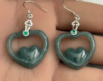 Può includere: Un paio di orecchini in Giada Blu Acqua di Grado A. Ogni orecchino presenta un design a cuore intagliato di colore verde acqua, con una forma a cuore più piccola intagliata all'interno. Gli orecchini hanno ganci color argento e sono adornati con piccole gemme chiare e verdi.