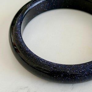 May include: A smooth, circular bangle bracelet crafted from dark blue sandstone, featuring a polished, reflective surface embedded with numerous tiny, glittering metallic flecks that create a starry, celestial appearance throughout the material.