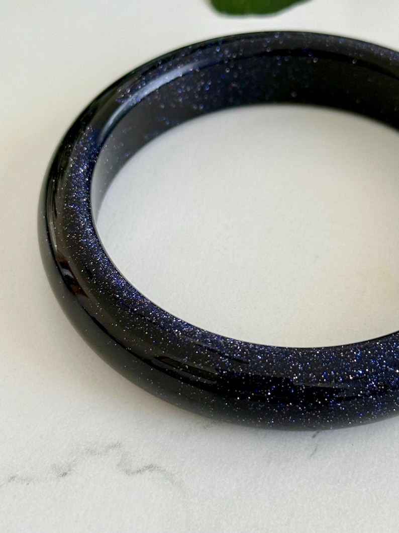 May include: A smooth, circular bangle bracelet crafted from dark blue sandstone, featuring a polished, reflective surface embedded with numerous tiny, glittering metallic flecks that create a starry, celestial appearance throughout the material.