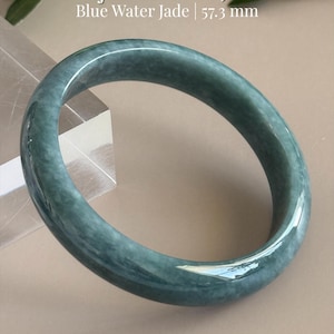 May include: A smooth, polished blue water jadeite bangle bracelet with a mottled teal and grey appearance. The text Certified Grade A Jadeite Blue Water Jade | 57.3 mm is visible above the bangle, and AmorGemsBoutique is printed at the bottom.