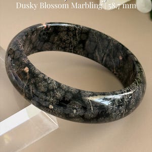 May include: A polished plum jade bangle with a dark, dusky blossom marbling pattern. The text on the image reads: Plum Jade Bangle, Dusky Blossom Marbling, 58.7 mm, and AmorGemsBoutique. The bangle has a smooth, rounded surface and a deep color.