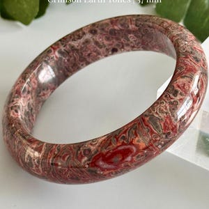 May include: A polished Leopard Skin Jasper bracelet with crimson earth tones. The bracelet is 57 mm in diameter and features a unique pattern of red, brown, and gray swirls. The text "Amor Gems Boutique" is at the bottom.