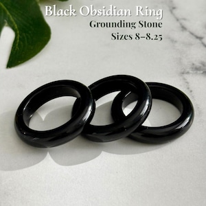 May include: Three black obsidian rings, sizes 8-8.25, are displayed. The rings are smooth and glossy, reflecting light. The text "Black Obsidian Ring, Grounding Stone" is visible. The rings are from AmorGems Boutique.