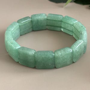 Pu&ograve; includere: Un braccialetto di avventurina verde. Il braccialetto &egrave; composto da perline rettangolari e lucide. Le perline sono di colore verde chiaro e hanno un aspetto leggermente traslucido. Il braccialetto &egrave; esposto su uno sfondo neutro.