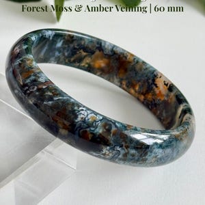 May include: A polished moss agate bangle featuring forest moss green and amber veining with a 60 mm diameter. The text on the image reads Moss Agate Bangle, Forest Moss & Amber Veining | 60 mm, and AmorGemsBoutique. The bangle has a smooth, rounded surface.