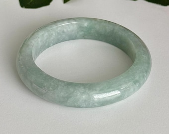 May include: A certified Grade A jadeite bangle in an icy celadon green color. The smooth, rounded bracelet features a natural Sajin pattern and measures 58 mm in diameter. The text Certified Grade A Jadeite, Icy Celadon Green, Natural Sajin, 58 mm, and AmorGemsBoutique is visible.