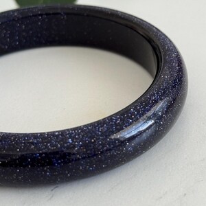 May include: A smooth, circular bangle bracelet crafted from dark blue sandstone, featuring a polished finish with numerous tiny, reflective silver-toned glitter particles embedded throughout the material, creating a starry night sky appearance.