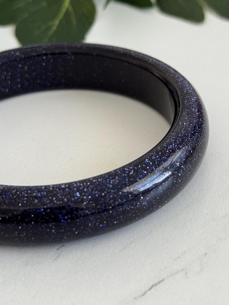 May include: A smooth, circular bangle bracelet crafted from dark blue sandstone, featuring a polished finish with numerous tiny, reflective silver-toned glitter particles embedded throughout the material, creating a starry night sky appearance.