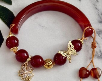 Può includere: Un bracciale a mezza luna in cornalina rossa lucida. Il bracciale presenta dettagli dorati, tra cui un ciondolo a farfalla, un pendente a fiore e perline rotonde. Il design regolabile include un cordino con due perline di cornalina.