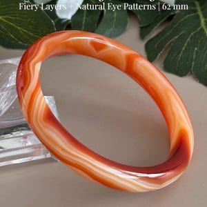 May include: A smooth, polished banded agate bangle featuring fiery orange and deep red layers with natural eye patterns. The bangle has a diameter of 62 mm. Text on the image reads: Banded Agate Bangle, Fiery Layers + Natural Eye Patterns | 62 mm, AmorGemsBoutique.
