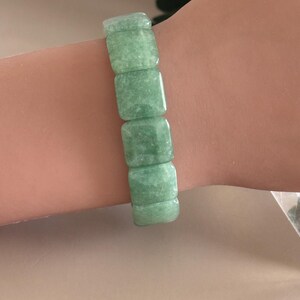 Pu&ograve; includere: Un braccialetto di giada verde chiaro. Il braccialetto &egrave; composto da pietre di giada rettangolari e lucide. Le pietre sono di un colore verde tenue e smorzato. Il braccialetto &egrave; indossato su un polso.