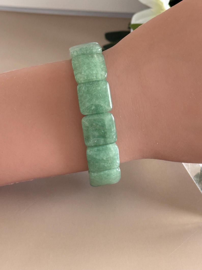 Pu&ograve; includere: Un braccialetto di giada verde chiaro. Il braccialetto &egrave; composto da pietre di giada rettangolari e lucide. Le pietre sono di un colore verde tenue e smorzato. Il braccialetto &egrave; indossato su un polso.