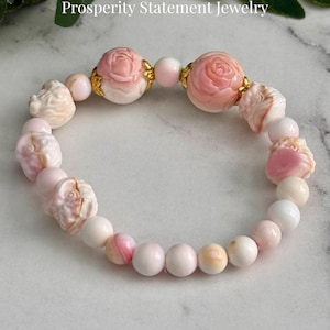 May include: A pink and white bracelet with carved conch shell beads and rose-shaped accents. The bracelet includes the text "Queen Conch Lotus Dragon Bracelet Prosperity Statement Jewelry" and "AmorGems Boutique".