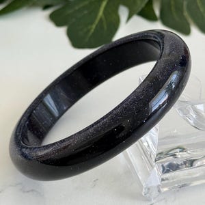 May include: A smooth, rounded bangle bracelet crafted from dark blue sandstone, featuring a polished finish with embedded glittery particles that create a starry, reflective appearance throughout the entire surface of the circular jewelry piece.