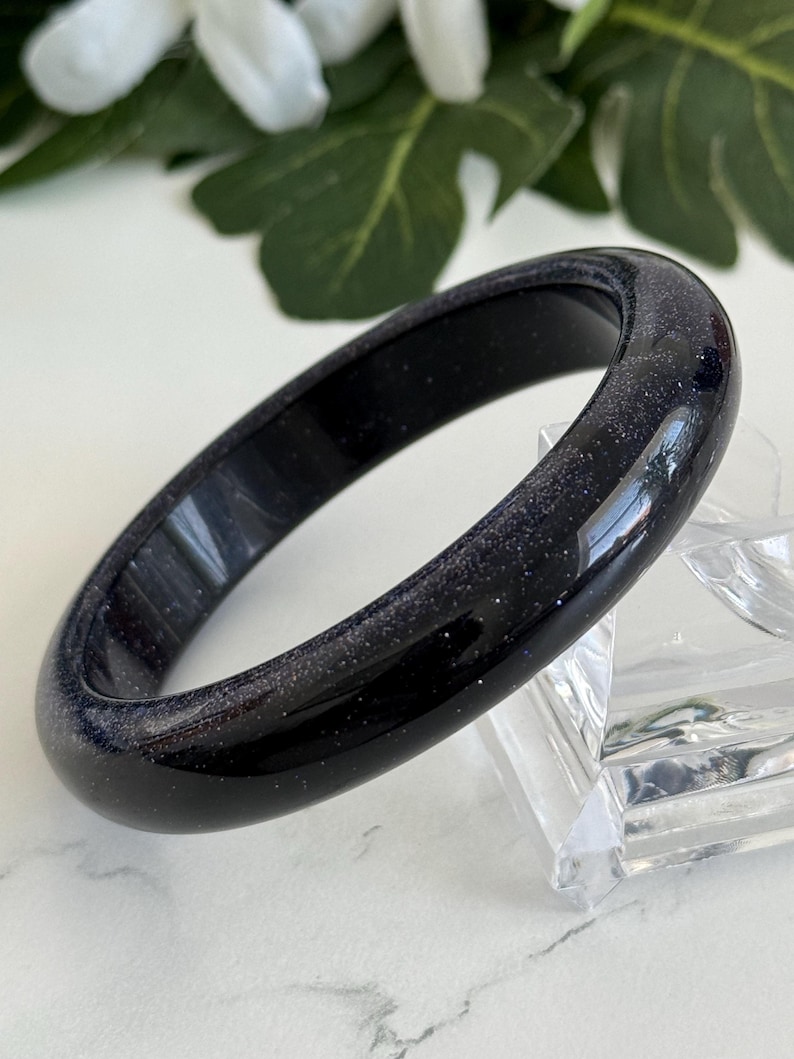 May include: A smooth, rounded bangle bracelet crafted from dark blue sandstone, featuring a polished finish with embedded glittery particles that create a starry, reflective appearance throughout the entire surface of the circular jewelry piece.