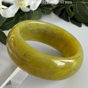 May include: A smooth, polished avocado green Xiuyan jade bangle with a 61 mm diameter. The text Natural Jade Bangle, Xiuyan | Avocado Green | 61 mm is visible above the bangle, and AmorGemsBoutique is printed at the bottom of the image.