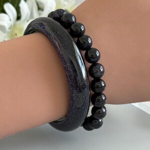 May include: A dark blue glittery bangle bracelet paired with a matching dark blue beaded stretch bracelet. Both pieces feature a deep navy hue with embedded sparkling flecks that catch the light, creating a starry night effect on the wrist.