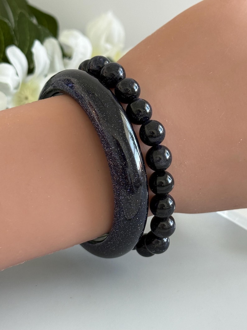 May include: A dark blue glittery bangle bracelet paired with a matching dark blue beaded stretch bracelet. Both pieces feature a deep navy hue with embedded sparkling flecks that catch the light, creating a starry night effect on the wrist.