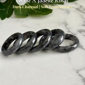 May include: Five dark charcoal jadeite rings with soft translucence are displayed. The rings are arranged in a row, showcasing their smooth, polished surfaces. The text "Grade A Jadeite Rings" and "Dark Charcoal | Soft Translucence" is visible.