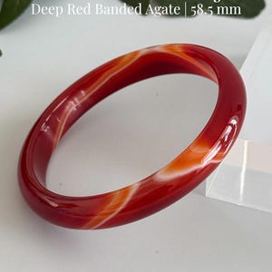 May include: A smooth, polished natural carnelian bangle featuring deep red and orange banded agate patterns. The bracelet has a 58.5 mm inner diameter. Text on the image reads: Natural Carnelian Bangle, Deep Red Banded Agate | 58.5 mm, AmorGemsBoutique.