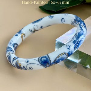 May include: Hand-painted porcelain butterfly bangle. The white bangle features blue butterfly and floral designs. The text on the image reads "Porcelain Butterfly Bangle", "Hand-Painted | 60-61 mm", and "AmorGems Boutique™".