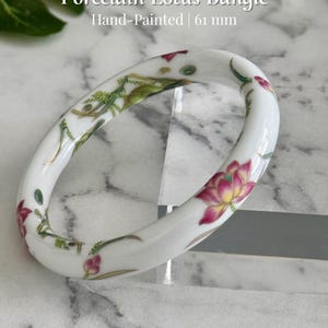 May include: Hand-painted porcelain lotus bangle. The white bangle features a floral design with pink and yellow lotus flowers and green leaves. The bangle is 61 mm in diameter.