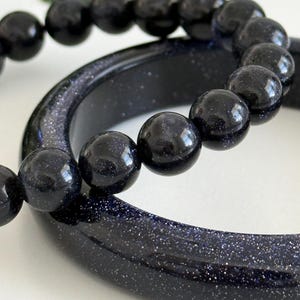 May include: A dark blue sandstone bangle bracelet with a smooth, polished surface and a matching beaded bracelet featuring round, dark blue sandstone beads with a shimmering, glitter-like appearance throughout the material.