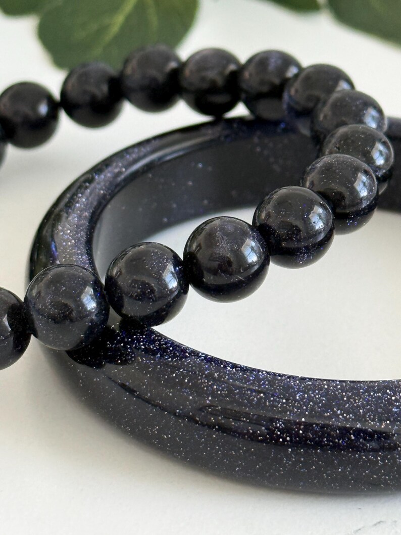 May include: A dark blue sandstone bangle bracelet with a smooth, polished surface and a matching beaded bracelet featuring round, dark blue sandstone beads with a shimmering, glitter-like appearance throughout the material.