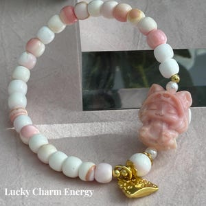 May include: A Queen Conch Lucky Cat bracelet with white, pink, and peach-colored beads. The bracelet features a carved pink cat charm and a gold-colored charm. The text "Lucky Charm Energy" is visible.