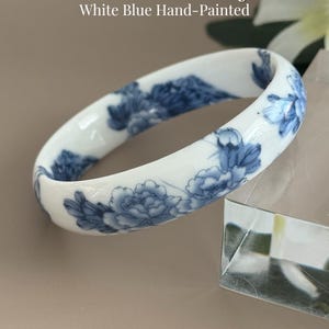 May include: A white porcelain bangle featuring a hand-painted blue floral pattern. The text "Porcelain Floral Bangle White Blue Hand-Painted" and "AmorGemsBoutique" are visible in the image. The bangle has a smooth, glossy finish and a rounded shape.