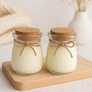 Peut inclure: Deux petits pots en verre avec des couvercles en liège et des nœuds en ficelle, remplis d'une substance blanche crémeuse. Les pots sont posés sur une planche en bois clair, suggérant un produit de spa ou de parfum d'intérieur.