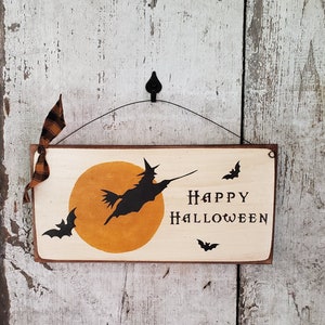 Może przedstawiać: Drewniany znak z czarną sylwetką czarownicy latającej na miotle przed ż&oacute;łtym księżycem. Znak głosi "Happy Halloween" i ma czarne nietoperze wok&oacute;ł krawędzi.