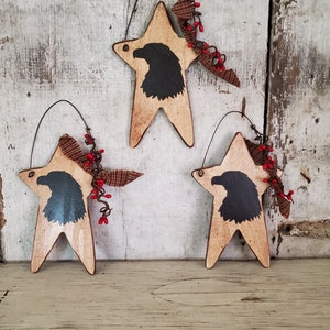 Puede incluir: Tres adornos de estrella de madera con una silueta negra de un &aacute;guila en cada uno. Las estrellas est&aacute;n pintadas en un acabado blanco y marr&oacute;n desgastado. Cada estrella tiene un lazo de tela rojo y blanco y ramitas de bayas rojas.