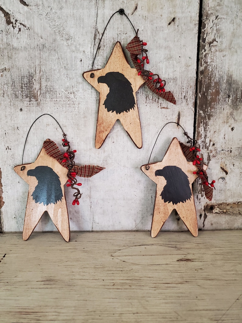 Puede incluir: Tres adornos de estrella de madera con una silueta negra de un &aacute;guila en cada uno. Las estrellas est&aacute;n pintadas en un acabado blanco y marr&oacute;n desgastado. Cada estrella tiene un lazo de tela rojo y blanco y ramitas de bayas rojas.