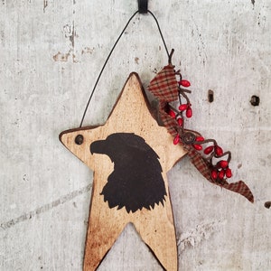 Puede incluir: Un adorno de madera en forma de estrella con una silueta negra de un &aacute;guila en el centro. La estrella est&aacute; pintada de color marr&oacute;n claro y tiene un aspecto desgastado. El adorno est&aacute; decorado con una cinta de cuadros rojos y blancos y bayas rojas.
