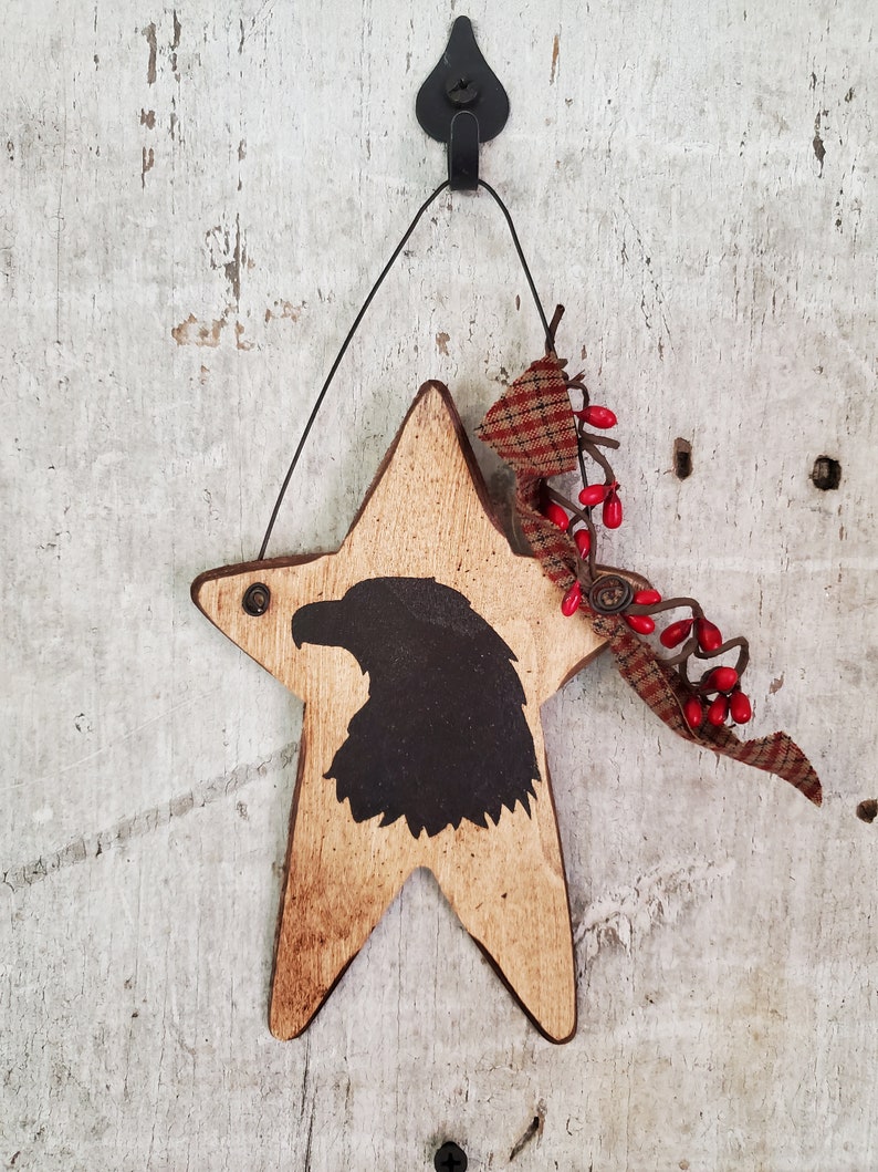 Puede incluir: Un adorno de madera en forma de estrella con una silueta negra de un &aacute;guila en el centro. La estrella est&aacute; pintada de color marr&oacute;n claro y tiene un aspecto desgastado. El adorno est&aacute; decorado con una cinta de cuadros rojos y blancos y bayas rojas.