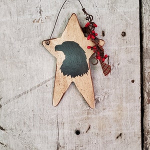 Puede incluir: Un adorno de madera en forma de estrella con una silueta pintada de la cabeza de un &aacute;guila. La estrella est&aacute; pintada de color marr&oacute;n claro y tiene un acabado desgastado. El &aacute;guila est&aacute; pintada de negro y tiene un tono verde. El adorno est&aacute; colgado de un lazo de alambre y tiene una peque&ntilde;a rama de bayas rojas unida a &eacute;l.